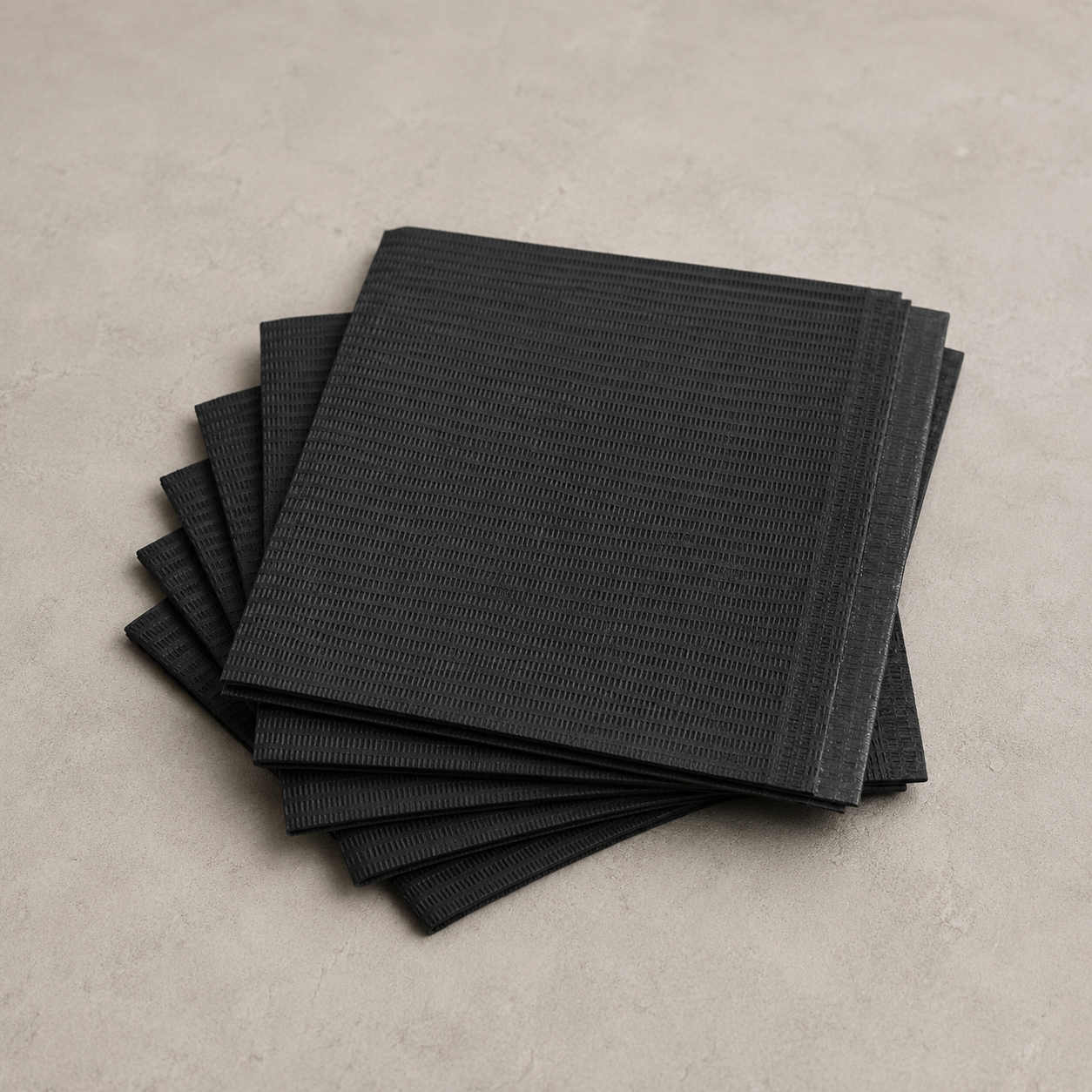 A stack of black textured paper napkins with a ribbed pattern, arranged on a neutral gray concrete-like surface.
Black Disposable Patient Bibs 125 PCS – Protective Tattoo Workstation Bibs