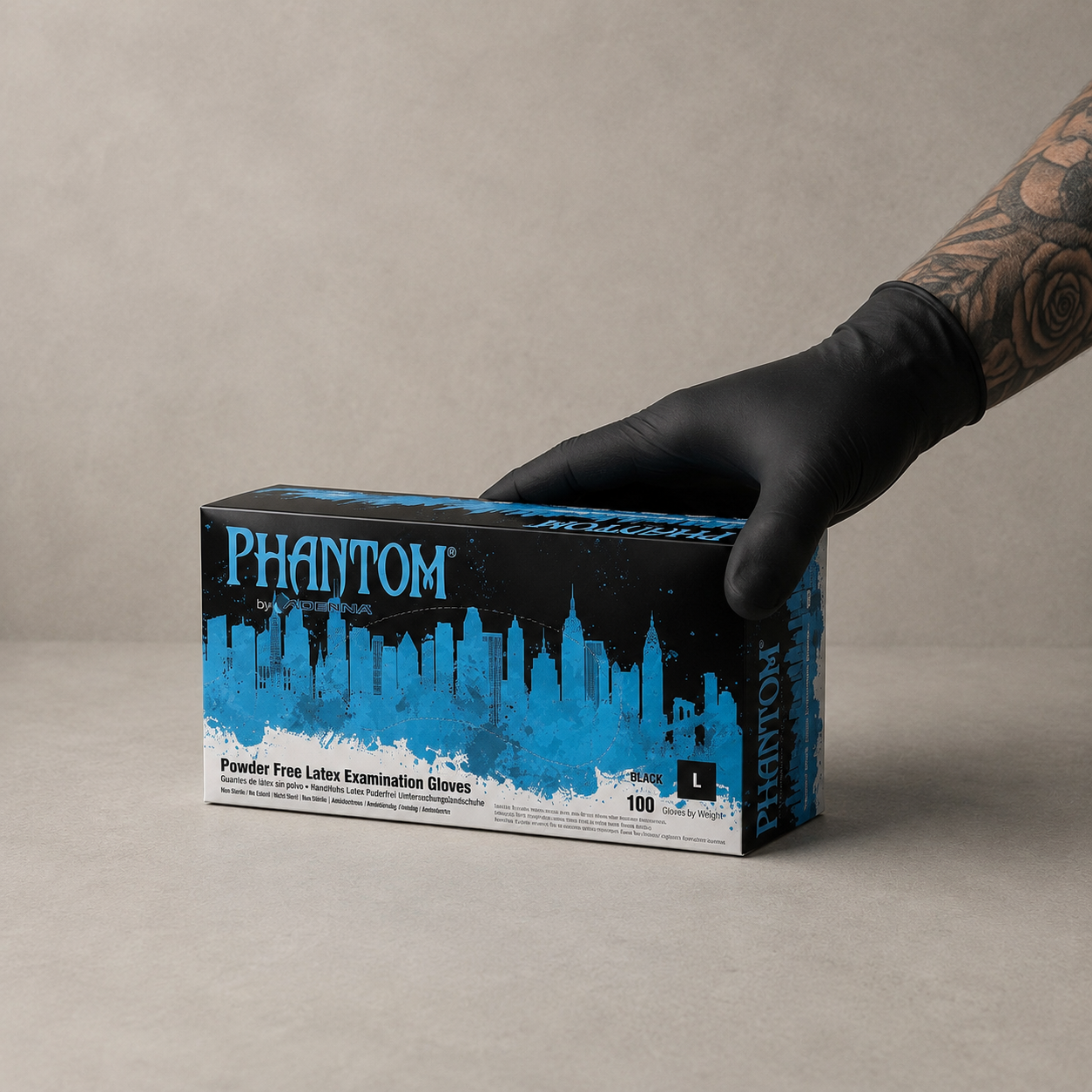 A hand wearing a black nitrile glove holds a box of Phantom Powder Free Latex Examination Gloves. The box is black with a bright blue city skyline graphic and white text, set against a neutral gray background.